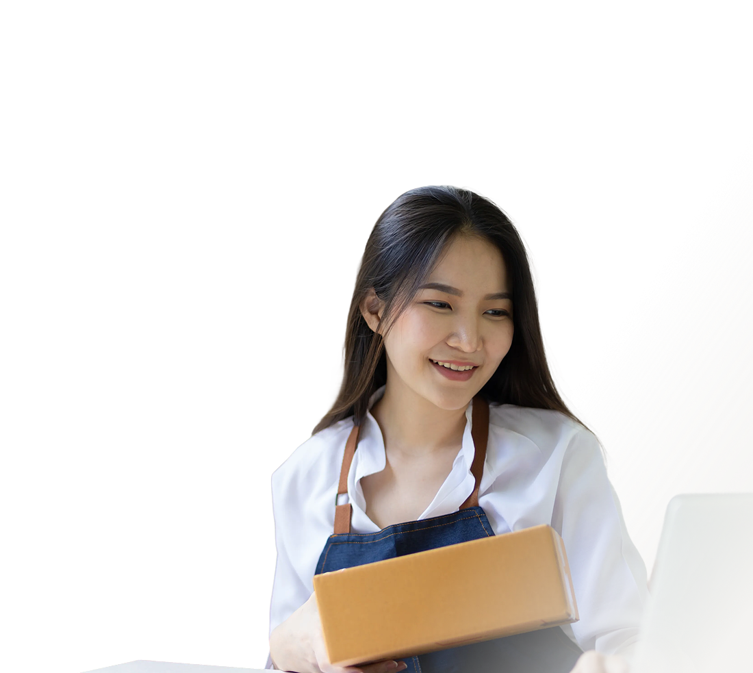 Smiling woman in a white shirt and blue apron holding a cardboard box and looking at a laptop.